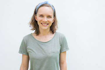 Happy cheerful woman in hairband and t-shirt looking at camera, smiling. Person posing isolated against white wall. Front view. Summer tourist outfit concept