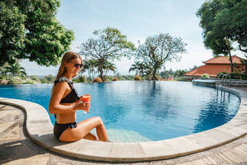 Woman swims to the edge of swimming pool, takes fresh juice, drinks it and swims away, sunny day in...