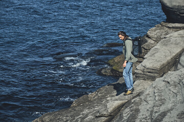 young long-haired man with a backpack and a photo tripod explores the rocky sea coast. Travel and outdoor concept.