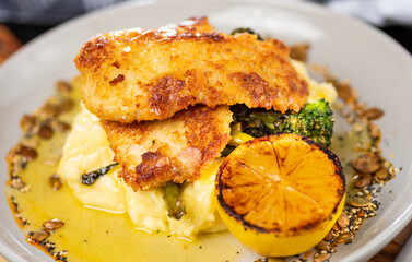 A serving of Chicken Schnitzel on a white plate with mashed potato, broccoli and lemon wedge. Side view close