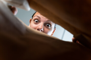 Caucasian woman peeking into craft paper delivery package with surprise emotion.