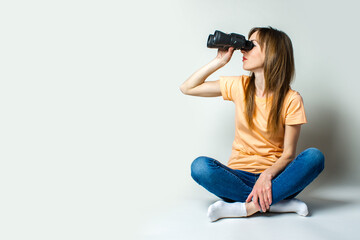 Young woman in a T-shirt and jeans sitting on the floor looks through binoculars on a light background. Banner