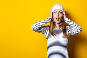 Naklejka premium Young woman surprised in shock holding hands to her head in a striped sweater and hat on a yellow background