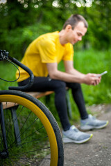 Obraz premium A young man sitting on a bench in a public Park communicates via mobile communication. In the background with his bike. Among trees and vegetation.