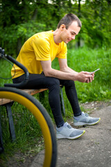 Obraz premium A young man sitting on a bench in a public Park communicates via mobile communication. In the background with his bike. Among trees and vegetation.