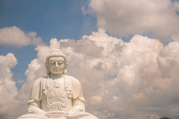 the solemn white statue of Buddha sitting on a high pedestal behind is a clear blue sky with white clouds