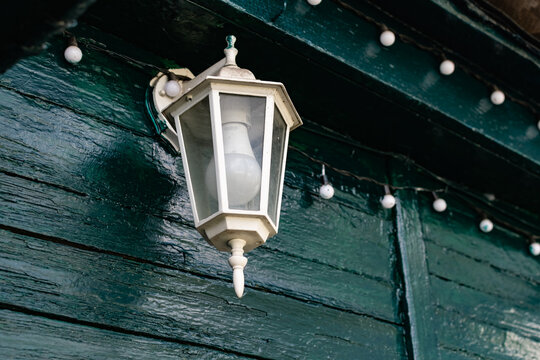 A white street lamp with its bulb turned off, hanging on a dark green wooden crowbar, next to long garlands with white round bulbs.