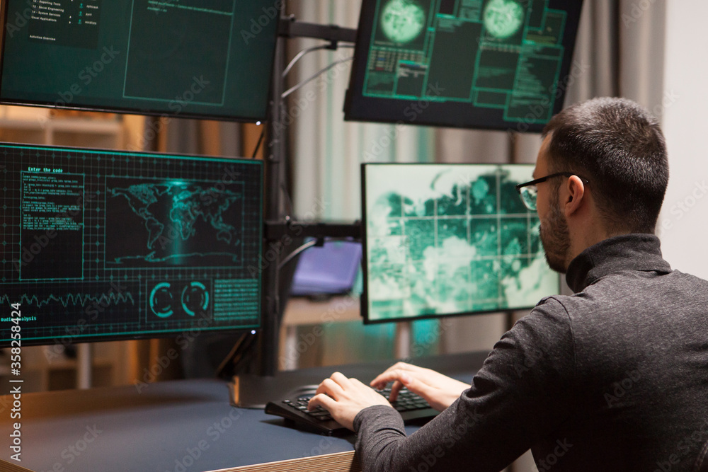 Poster young hacker with glasses writing a dangerous virus on computer with multiple screens.