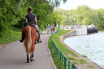 Girl riding horse in a summer park. Horsewoman on river coast on walking people background