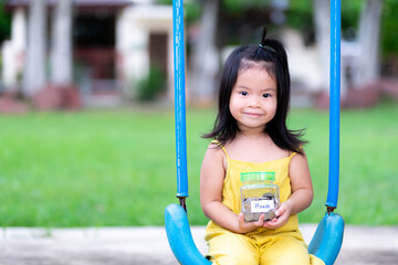 Children holding glass jars that put money coins for saving. At the jar was a message on white piece of paper that house. Cute Asian girls wear yellow outfits to sit on swing. Sweet smile on fresh air