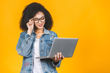Young african american black positive cool lady with curly hair using laptop and smiling isolated over yellow background.