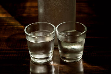 Frozen glasses with alcohol near the bottle. Drinks on a dark brown table. Transparent utensils on a black background. Old shabby boards under glass.