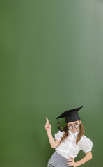 Little girl wearing graduation cap stands near empty green chalkboard and points up on empty space. Idea concept. Empty space for text