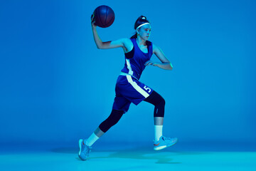 Plakat Desire to win. Young caucasian female basketball player training, prcticing with ball isolated on blue background in neon light. Concept of sport, movement, energy and dynamic, healthy lifestyle.