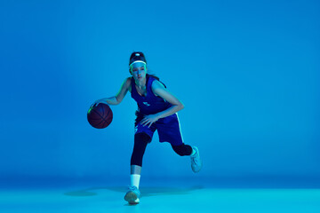 Desire to win. Young caucasian female basketball player training, prcticing with ball isolated on blue background in neon light. Concept of sport, movement, energy and dynamic, healthy lifestyle.