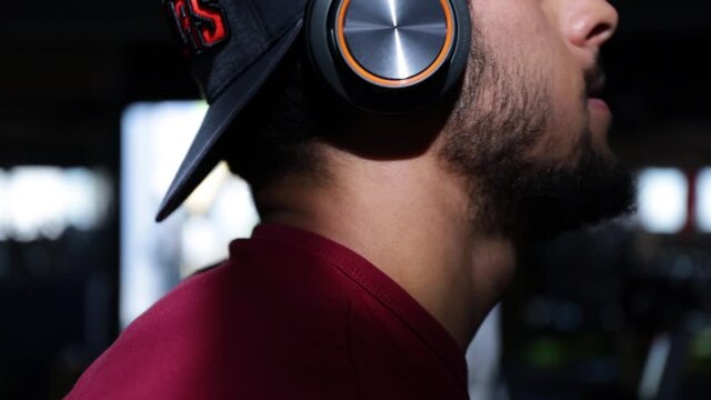 Young Caucasian bearded man wearing hat and headphones doing biceps workout with barbell in gym, close up profile
