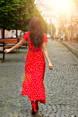 Happy girl walks down the street with cobblestones. Woman in a red dress with white spots. Dresses and hair are blown away by the wind. Girl on a background of blurred city street.