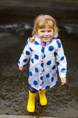 Little toddler girl wearing yellow rain boots, running and walking during sleet on rainy cloudy day. Cute happy child in colorful clothes jumping into puddle, splashing with water, outdoor activity