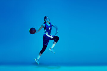 Fototapeta na wymiar In high flight. Young caucasian female basketball player training, prcticing with ball isolated on blue background in neon light. Concept of sport, movement, energy and dynamic, healthy lifestyle.
