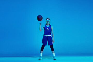 Young caucasian female basketball player of team posing confident with ball isolated on blue background in neon light. Concept of sport, movement, energy and dynamic, healthy lifestyle. Training.