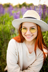 Young red haired girl with long hair in a wicker hat with a ribbon. Woman sitting with on a field of lupins at sunset and smiling