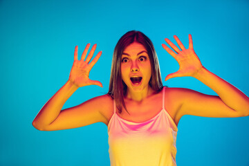 Astonished, surprise. Caucasian young woman's portrait on blue background in neon light. Beautiful female model in casual wear. Concept of human emotions, facial expression, sales, ad. Copyspace.