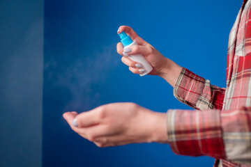 Spray disinfection, protection, prevention, COVID-19, coronavirus, safety, sanitation concept. Woman in red plaid shirt spraying antiseptic on hands against blue wall - close up, selective focus