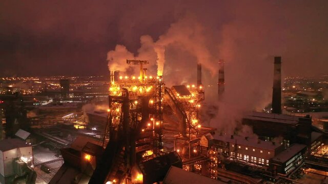 Aerial view of metallurgical plant blast furnace at night with smokestacks and fire blazing out of the pipe. Industrial panoramic landmark with blast furnance of metallurgical production