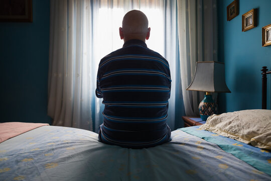 Back view of an old man sitting on his bed looking at the window
