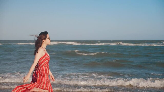 Slow Motion Of Asian Woman Walking Towards Sandy Ocean Beach Wearing A Long Flowing Dress With Sunset In The Distance On Summer Vacation.