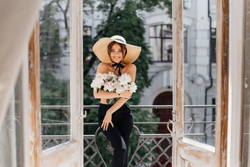 Stylish portrait of a girl on a vintage balcony who covers her breast with fresh peonies and on her head a stylish straw slag with a black ribbon around her face