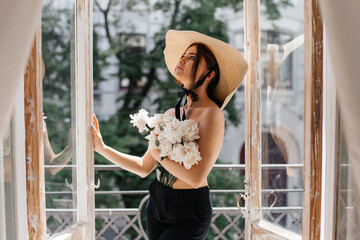 Stylish portrait of a girl on a vintage balcony who covers her breast with fresh peonies and on her head a stylish straw slag with a black ribbon around her face © nastya_shemet