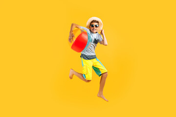 Fototapeta na wymiar Full-length portrait of a happy excited teenager in sunglasses and a summer hat, having fun and jumping, holding an inflatable sea ball on a yellow background