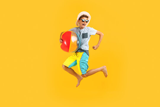 Full-length portrait of a happy excited teenager in sunglasses and a summer hat, having fun and jumping, holding an inflatable sea ball on a yellow background