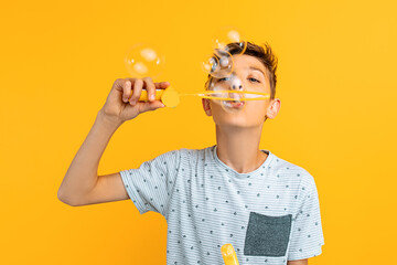 beautiful teen guy blows soap bubbles while standing on an isolated yellow background