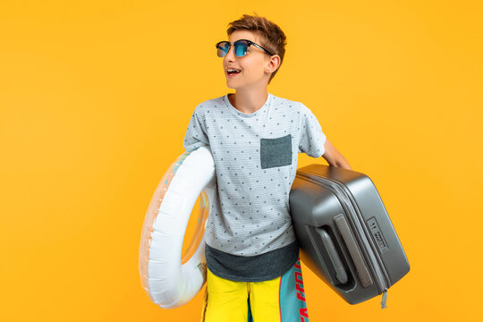 happy teenager in sunglasses stands with a suitcase and an inflatable sea circle on a yellow background. Concept of summer vacation, travel