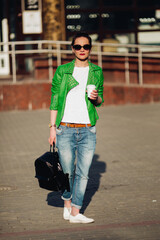 Stylish and beautiful brunette girl wearing in green leather jacket, jeans, white t shirt with cup of coffee and holding black bag.Fashionable woman walking at street after shopping. Sunny spring day.