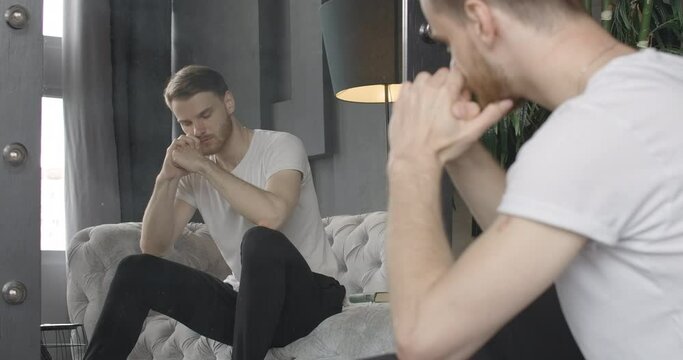 Reflection of unhappy young handsome man sighing and putting head on hands. Stressed Caucasian brunette guy reflecting in mirror indoors. Sadness, stress, problems. Cinema 4k ProRes HQ.