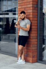 Portrait of handsome young man listening to music after running. Side view of young man in sportswear using his smart phone. Athlete man in earphones is having rest and listening to music. Brick wall.