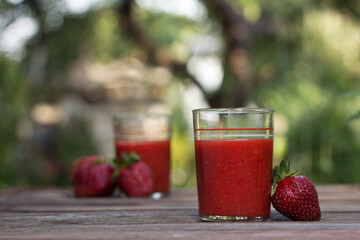 Strawberry juice with glass glasses on a wooden table and on the background of a green garden, on the table are ripe strawberries. Useful food, background, place for text.