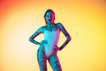 Confident. Young caucasian woman in swimsuit posing sensual on gradient yellow background in neon. Beautiful model with tattoos. Human emotions, sales, ad concept. Resort and vacation, summertime.