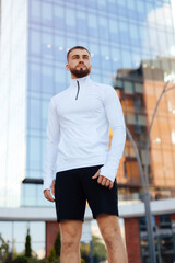 Cheerful male athlete resting after street workout session. Stylish urban young man in sportwear with a stylish hairstyle and beard stands near modern buildings in the city. Urban environment.