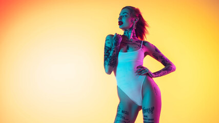 Young caucasian woman in swimsuit posing sensual on gradient yellow background in neon. Beautiful model with tattoos. Human emotions, sales, ad concept. Resort and vacation, summertime. Flyer.