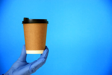 Hand in blue glove holds disposable cardboard cup of coffee on blue background. Safe take-out coffee or delivery service. Copy space.