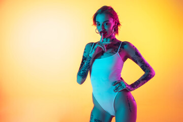 Sunlight. Young caucasian woman in swimsuit posing sensual on gradient yellow background in neon. Beautiful model with tattoos. Human emotions, sales, ad concept. Resort and vacation, summertime.