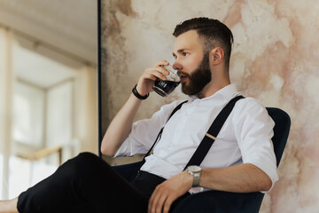 Successful businessman sitting in hotel  drinks whiskey. Serious man in business clothes working from hotel. Young handsome man relaxing at his apartment in a hotel after business meeting.