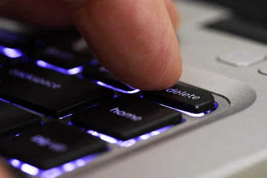 Extreme close up of a finger pressing the delete tab on a computer keyboard. Blue back lit computer laptop keys with a digit pushing the delete button.