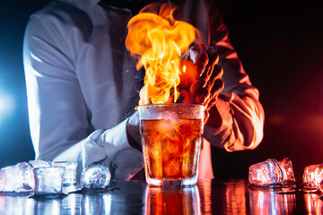 Barman making a fresh alcoholic cocktail with a smoky note on the dark bar counter and background.