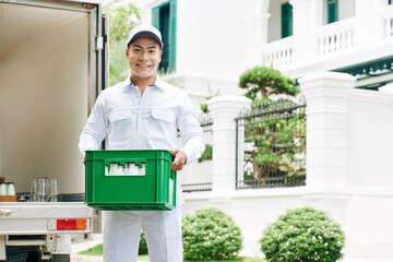 Horizontal medium long portrait shot of modern milkman wearing white clothes standing outdoors holding plastic box with milk bottles, copy space