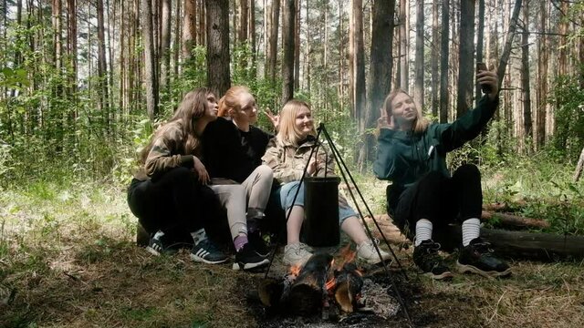 young girls sit by the fire in the forest. Fried sausages on a camping trip. They make tea. Active lifestyle in nature. summer trip. Friendship and leisure concept